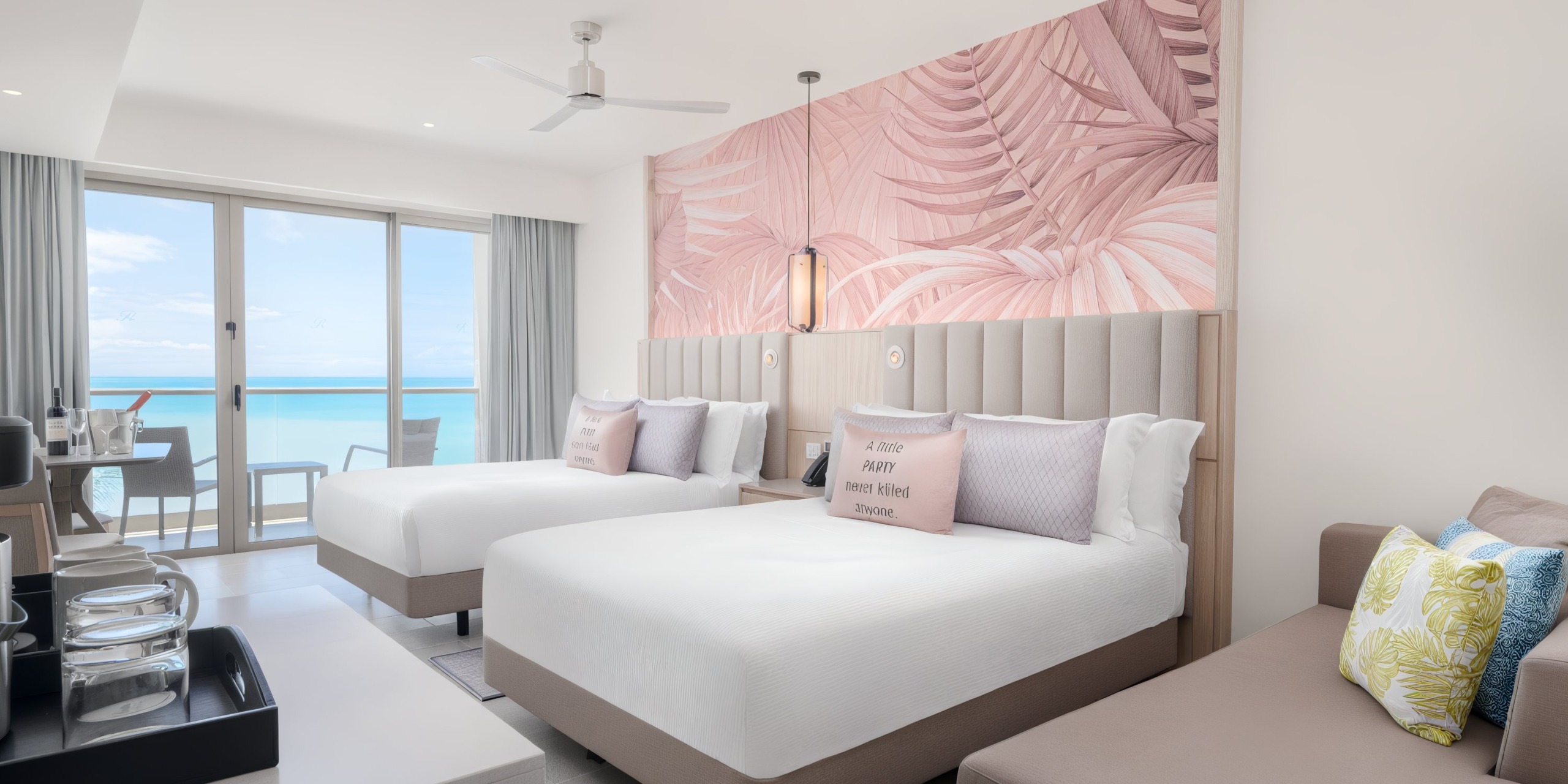 Royalton Chic Antigua offers a hotel room with two beds, a tropical-themed wall mural, a balcony with an ocean view, and a seating area adorned with decorative pillows.