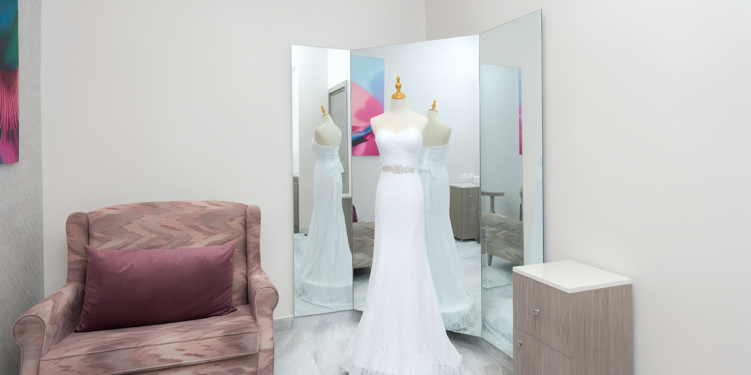 A Royalton Chic Antigua strapless white wedding dress is displayed on a mannequin and reflected in a three-sided mirror. To the left is a cushioned chair, and to the right is a small cabinet.