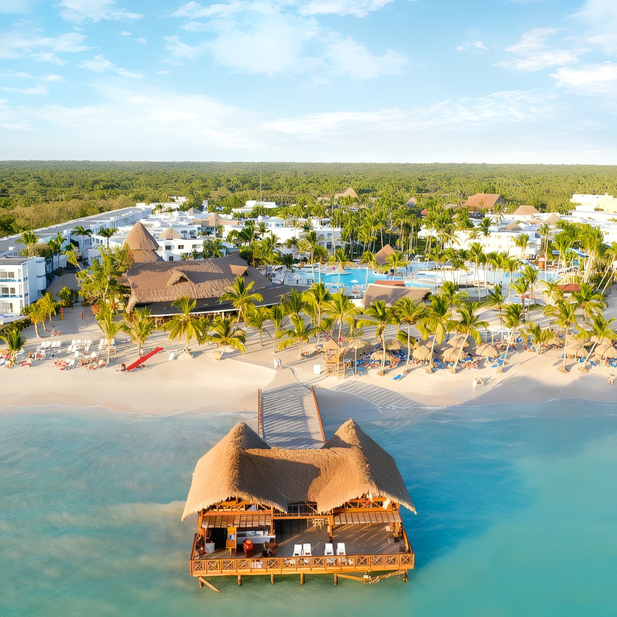 An aerial view of Sunscape Dominicus La Romana showcases a tropical beach with thatched-roof huts on the water, surrounded by palm trees and beachfront buildings.