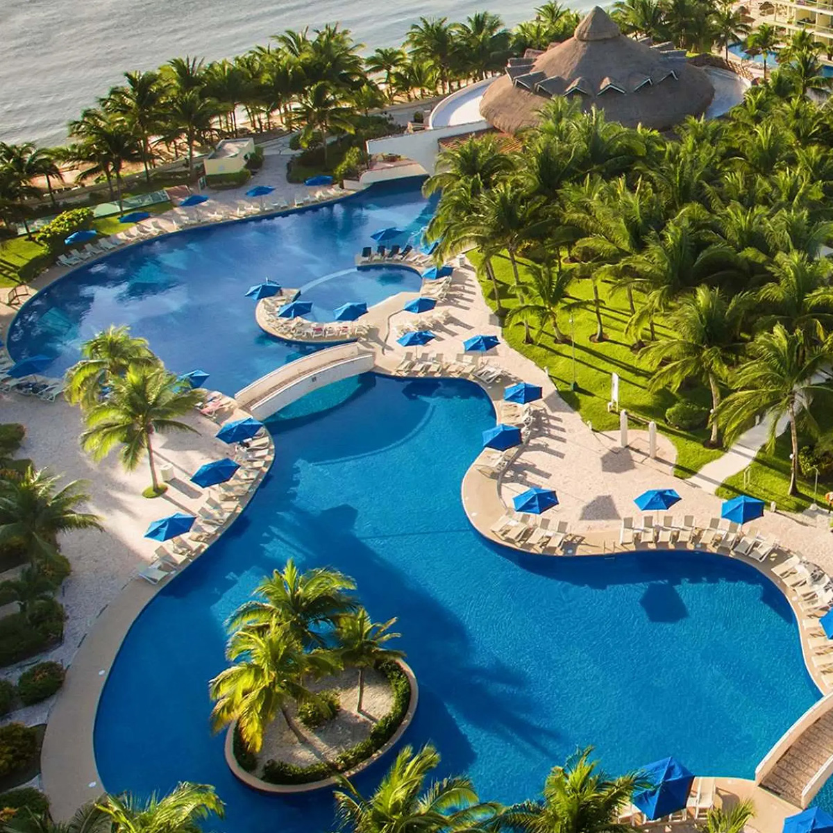 Aerial view of Azul Beach Resort Riviera Cancun featuring a sprawling, curvy blue pool, encircled by swaying palm trees and lounge chairs with blue umbrellas, positioned next to a thatched-roof building by the ocean.