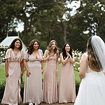 A bride in a white dress faces four bridesmaids in matching beige dresses, all standing outside in a garden setting.