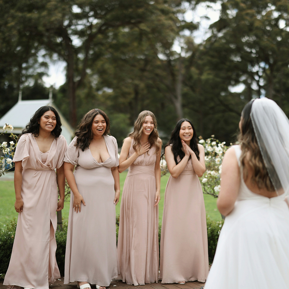 A bride in a white dress faces four bridesmaids in matching beige dresses, all standing outside in a garden setting.
