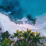 Aerial view of a tropical beach with clear blue water, white sand, palm trees, and small waves breaking near the shore.