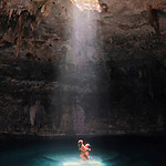 A person stands in a cave pool under a beam of sunlight streaming through an opening above.