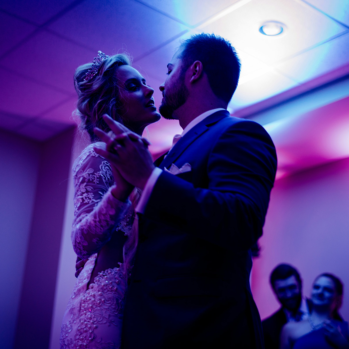 Bride and groom share a dance in a room with purple lighting, surrounded by a few guests.
