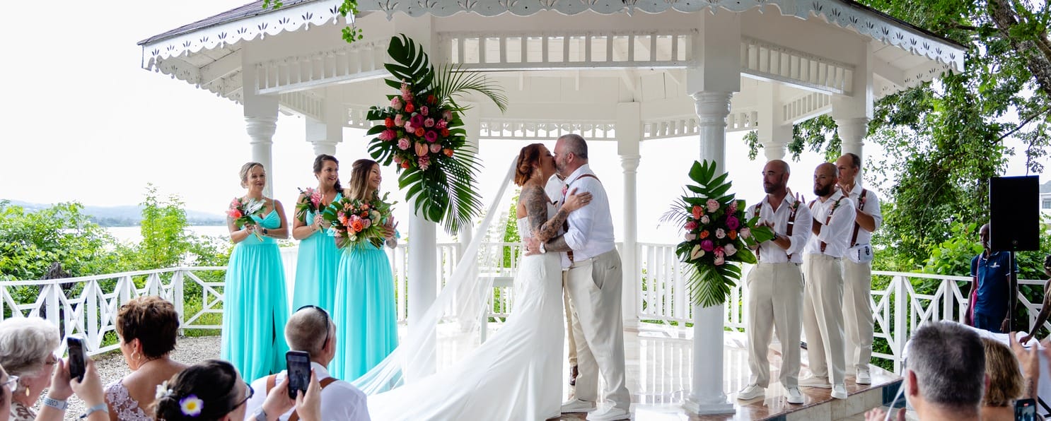 A couple kisses at their destination wedding in Jamaica beneath a white gazebo, guests watching.