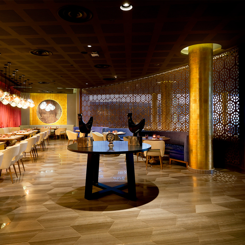 A dimly lit restaurant interior with modern decor, featuring a round table with sculptures, intricate wall patterns, and seating with cream-colored chairs under round hanging lights.