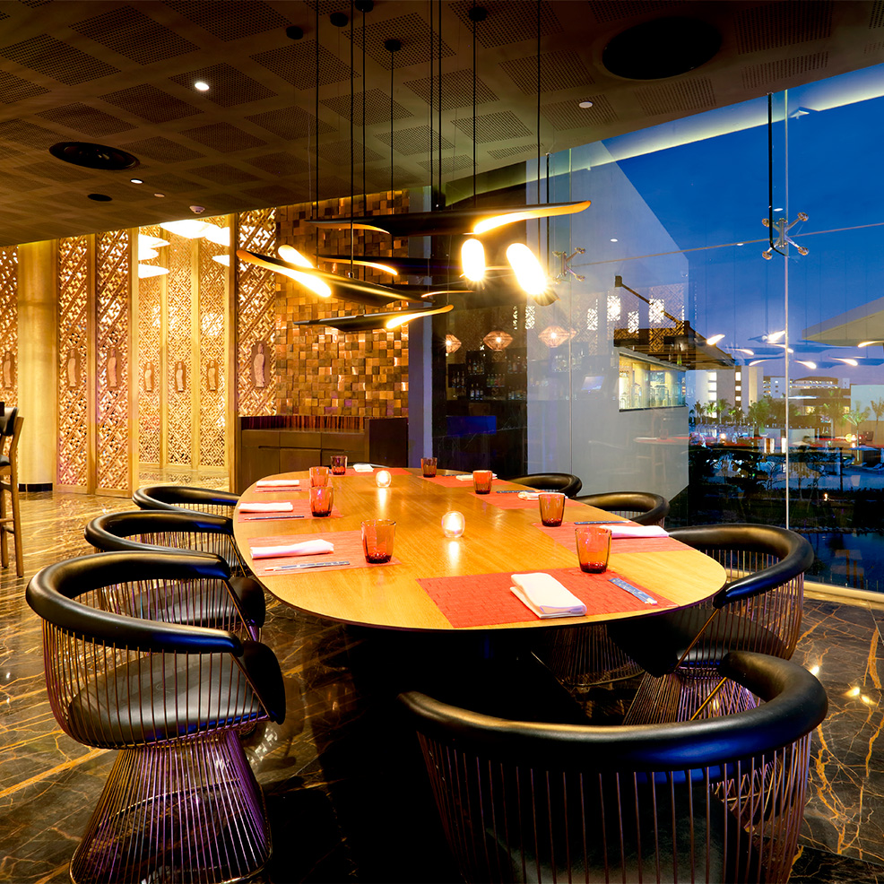 A modern dining room with a long oval table set for eight, surrounded by stylish chairs. The room features warm lighting, decorative walls, and large windows with a view of city lights.