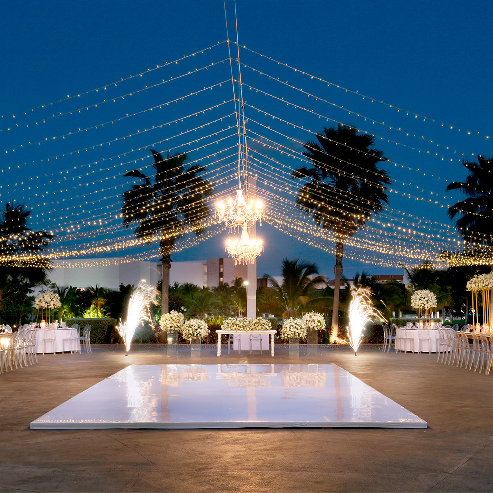 An outdoor event space with a white, glossy dance floor, surrounded by tables and chairs, under string lights and chandeliers, with fireworks and palm trees in the background.