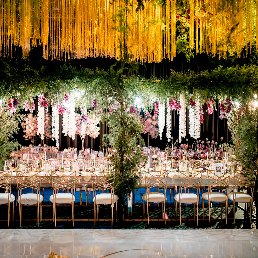 Elegant wedding reception hall with long tables set for dining. Overhead, a canopy of lush greenery, pink and white flowers, and golden streamers creates a festive atmosphere.