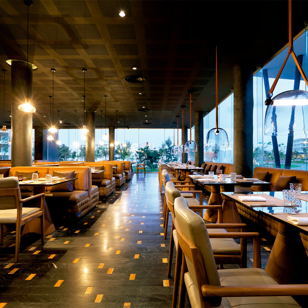 A modern restaurant interior with wooden tables, cushioned chairs, soft lighting, and large windows showcasing an outdoor view.