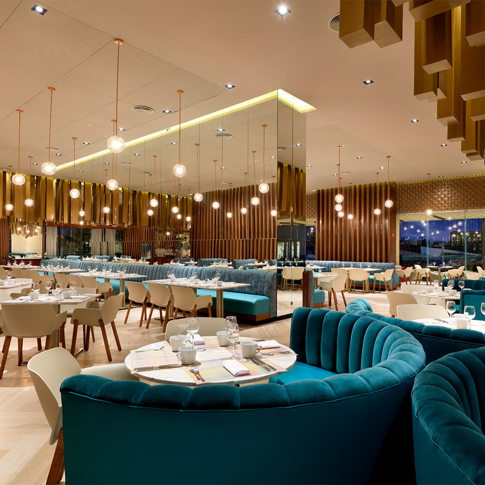 Elegant restaurant interior with teal velvet seating, wooden accents, and pendant lights. Tables are set for dining, and large windows overlook a night view.