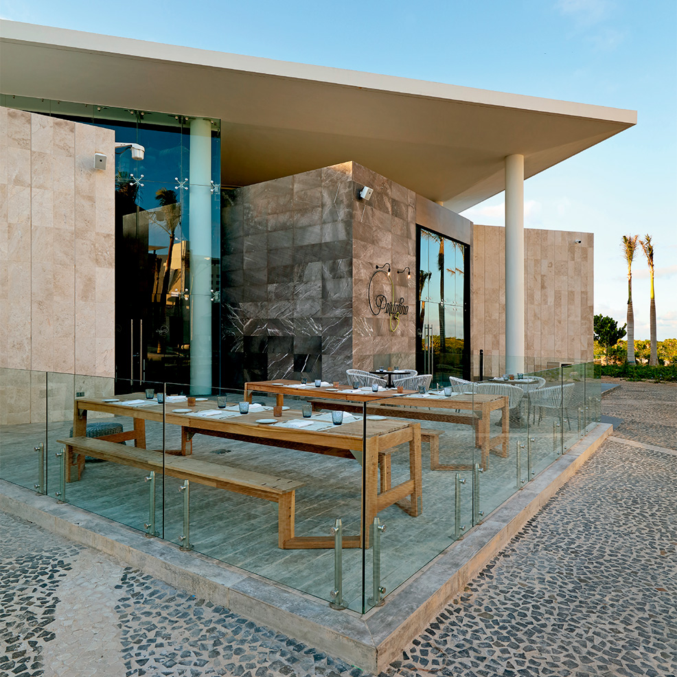 Modern outdoor seating area with wooden benches and tables, enclosed by glass panels. The building features large windows and a flat roof. Palm trees are visible in the background.