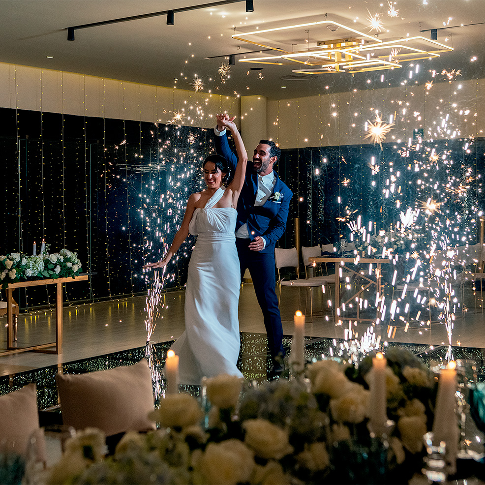 A bride and groom dance surrounded by sparkling fireworks on the floor, with elegant flower arrangements and dim lighting in the background.