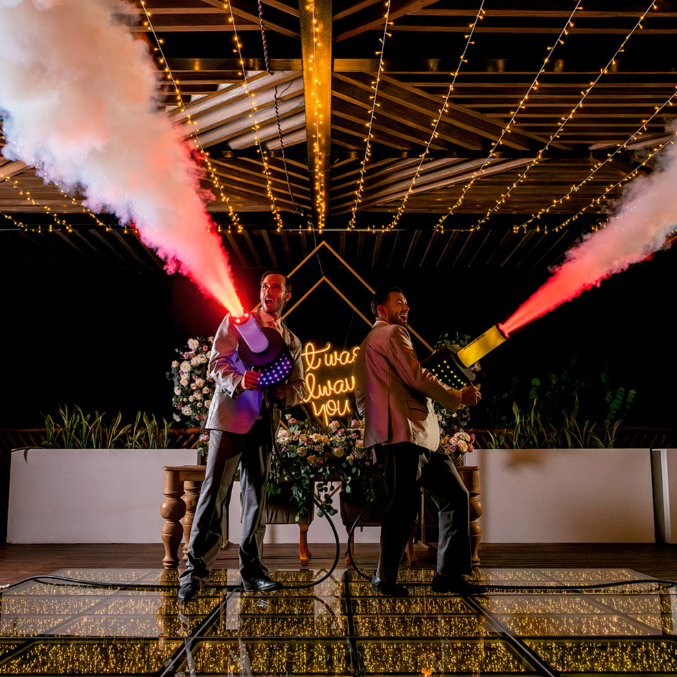 Two people in suits perform a special effect with smoke cannons emitting red smoke on a decorated stage with string lights and a neon sign.