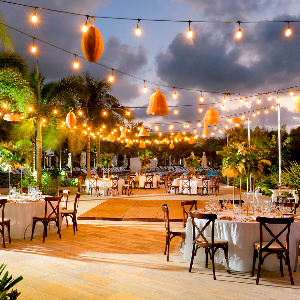 Outdoor dining area with round tables set for a meal, string lights overhead, and palm trees in the background against a cloudy evening sky.