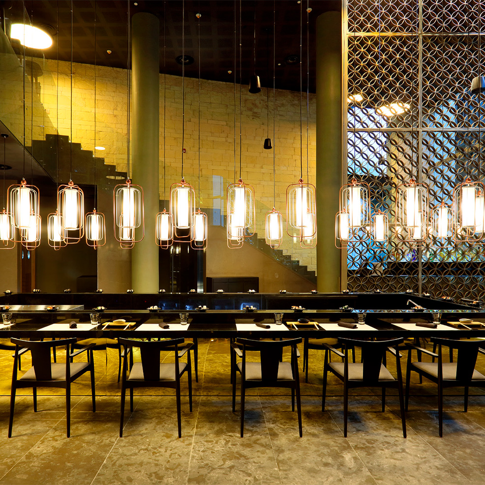 A modern restaurant interior with long pendant lights above a communal table. Stone walls and a decorative lattice window are in the background.