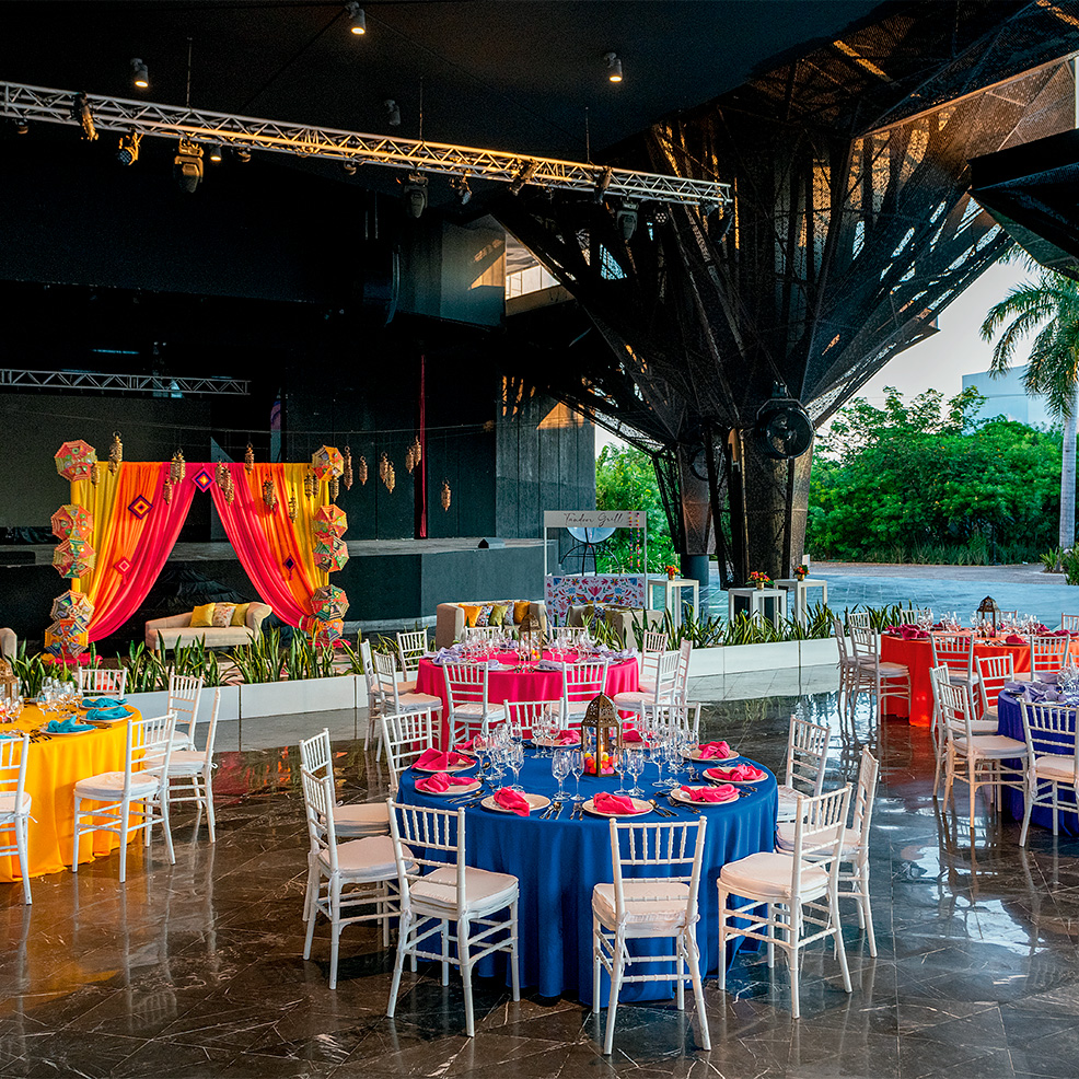 Colorful banquet setup with tables and chairs. A bright pink and yellow draped backdrop stands in the background. The venue features a modern design with lots of natural light.