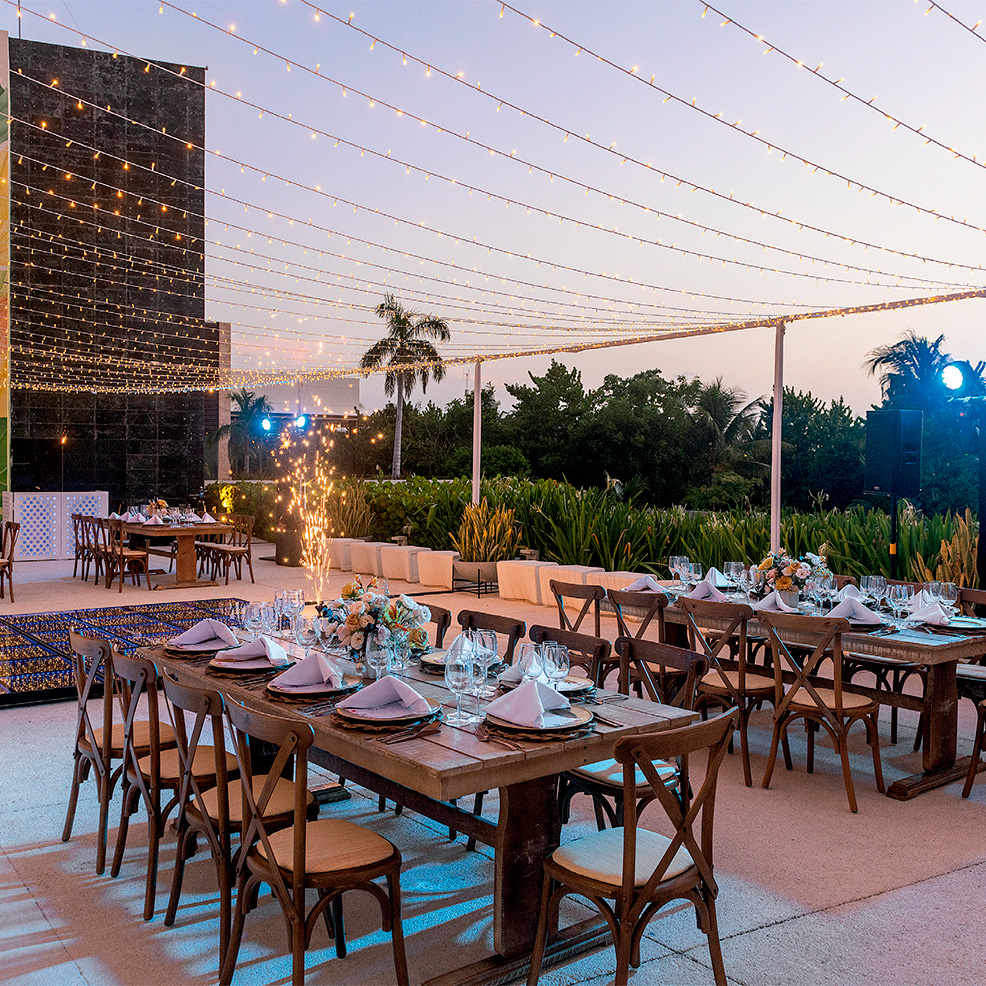 Outdoor dining area with wooden tables set with plates, napkins, and floral centerpieces. String lights are overhead, and greenery surrounds the space.