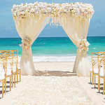 A beach wedding arch adorned with white and pink flowers, surrounded by elegant white chairs with golden frames, sets the stage for a stunning ceremony at weddings at Hard Rock Punta Cana against the breathtaking turquoise ocean backdrop.