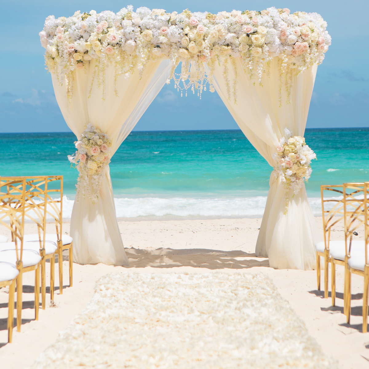 A beach wedding arch adorned with white and pink flowers, surrounded by elegant white chairs with golden frames, sets the stage for a stunning ceremony at weddings at Hard Rock Punta Cana against the breathtaking turquoise ocean backdrop.