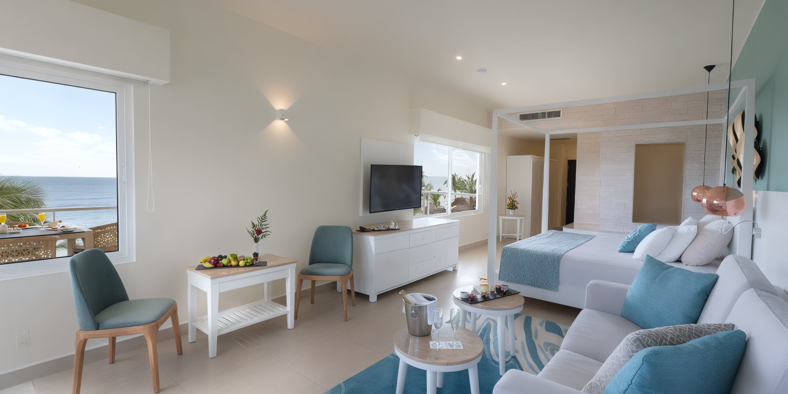 Enjoy a bright, modern room at Azul Beach Resort Riviera Cancun, featuring a spacious bed, sofa, TV, and a comfortable seating area. The window provides a picturesque ocean view. The room's decor showcases calming blue and white tones, complemented by a small dining table and an inviting fruit platter.