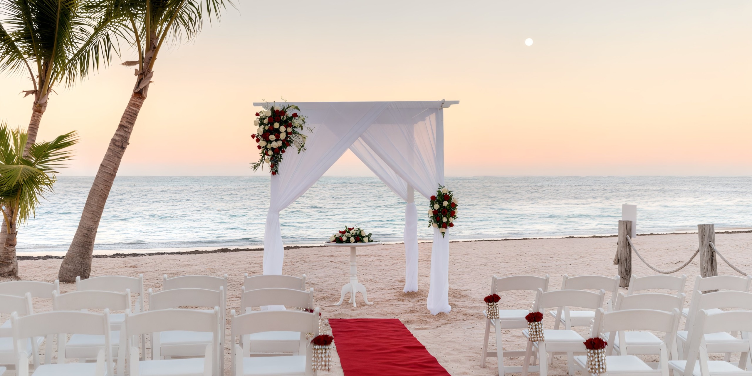 A beach wedding setup at Lopesan Costa Bavaro features a stunning white arch adorned with red flowers, a vibrant red aisle runner, and elegant rows of white chairs all oriented towards the ocean, beautifully framed by palm trees against the backdrop of a sunset sky.
