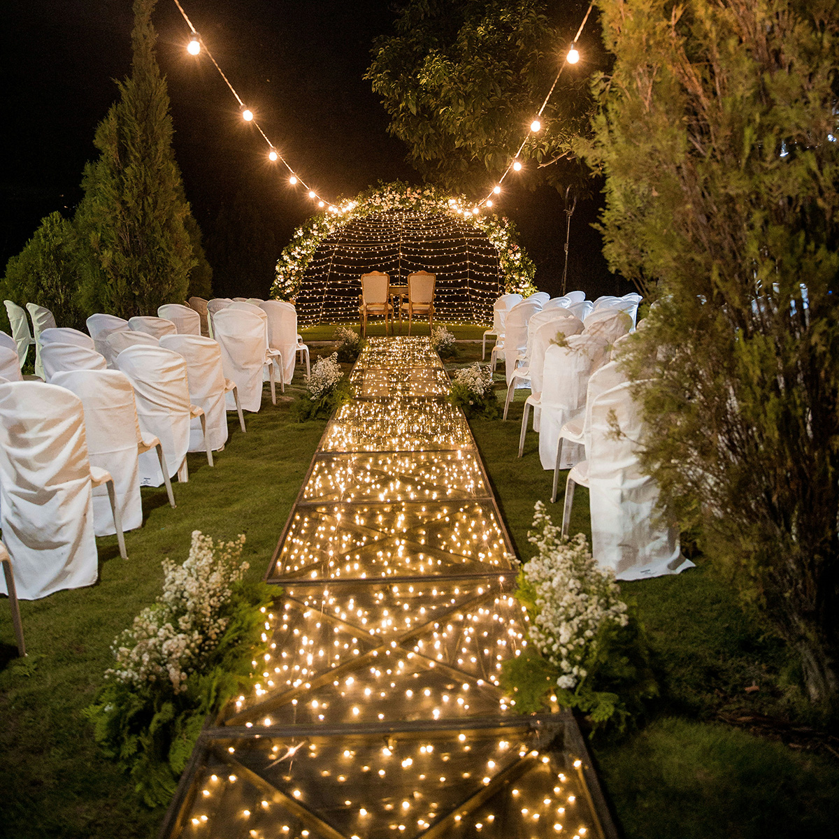 Outdoor wedding setup with a lighted aisle, white chair covers, and floral arrangements. A decorative arch with lights and two chairs is at the end of the aisle, surrounded by trees.