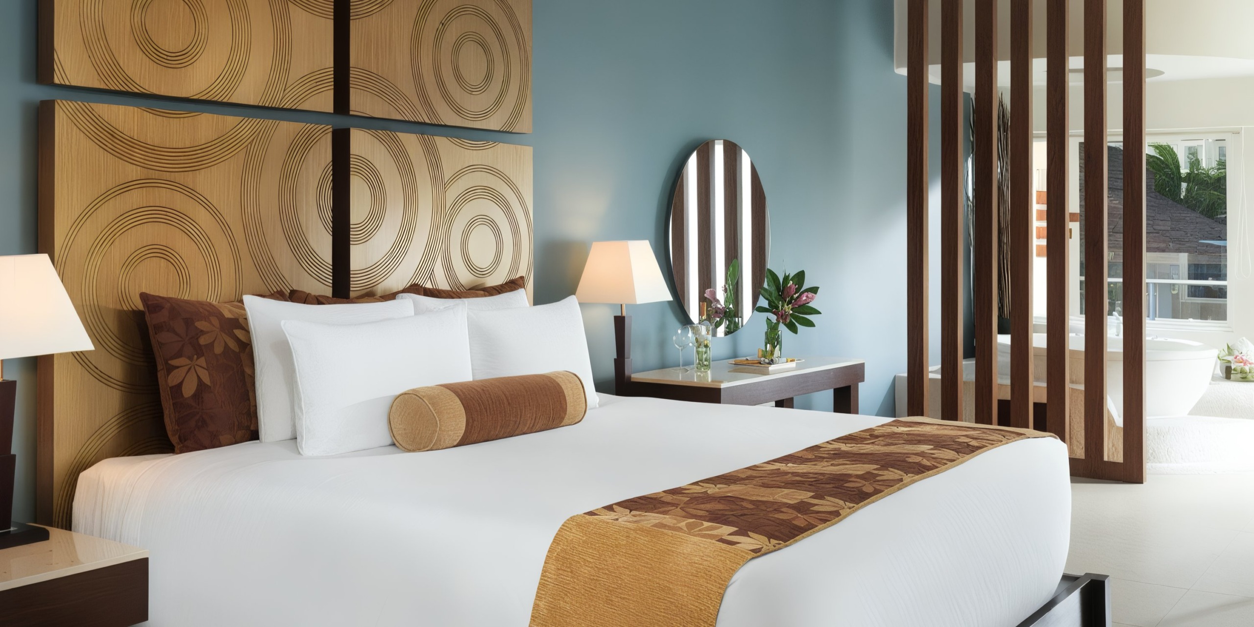 A contemporary room at the Azul Beach Resort Riviera Cancun featuring a bed, two bedside lamps, circular artwork, and a round mirror. Through wooden slats, there's a glimpse of a bathroom with a tub.