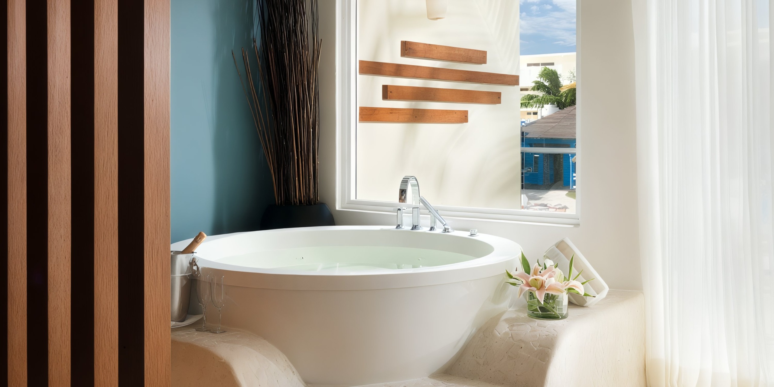 At the Azul Beach Resort Riviera Cancun, a round bathtub filled with water is positioned near a large window. Beside the tub, you'll find a champagne bottle nestled in an ice bucket and a vase adorned with flowers.