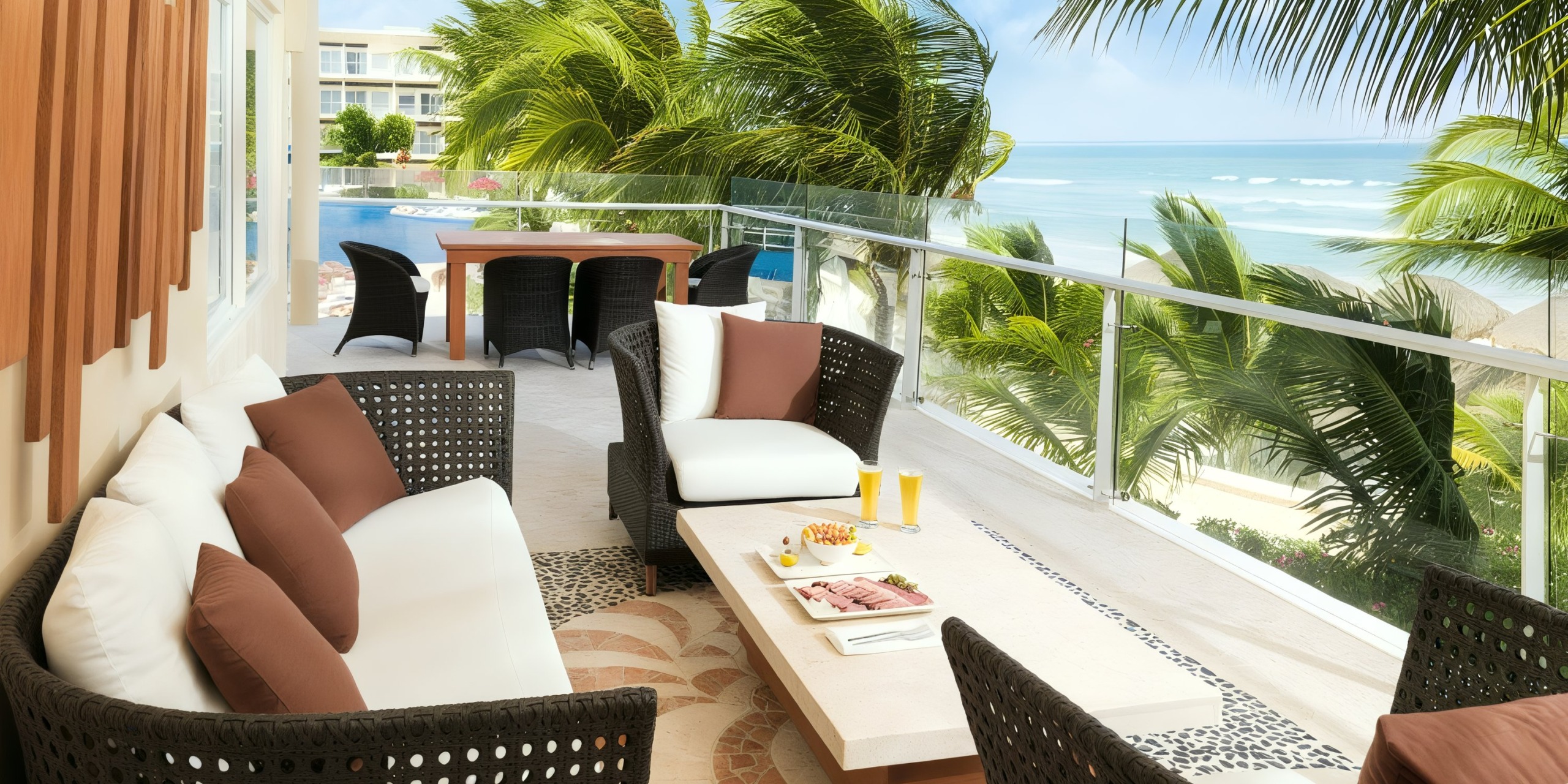 A patio at Azul Beach Resort Riviera Cancun features wicker furniture and a table with snacks and drinks, offering a view of the pool, beach, and palm trees in the background.