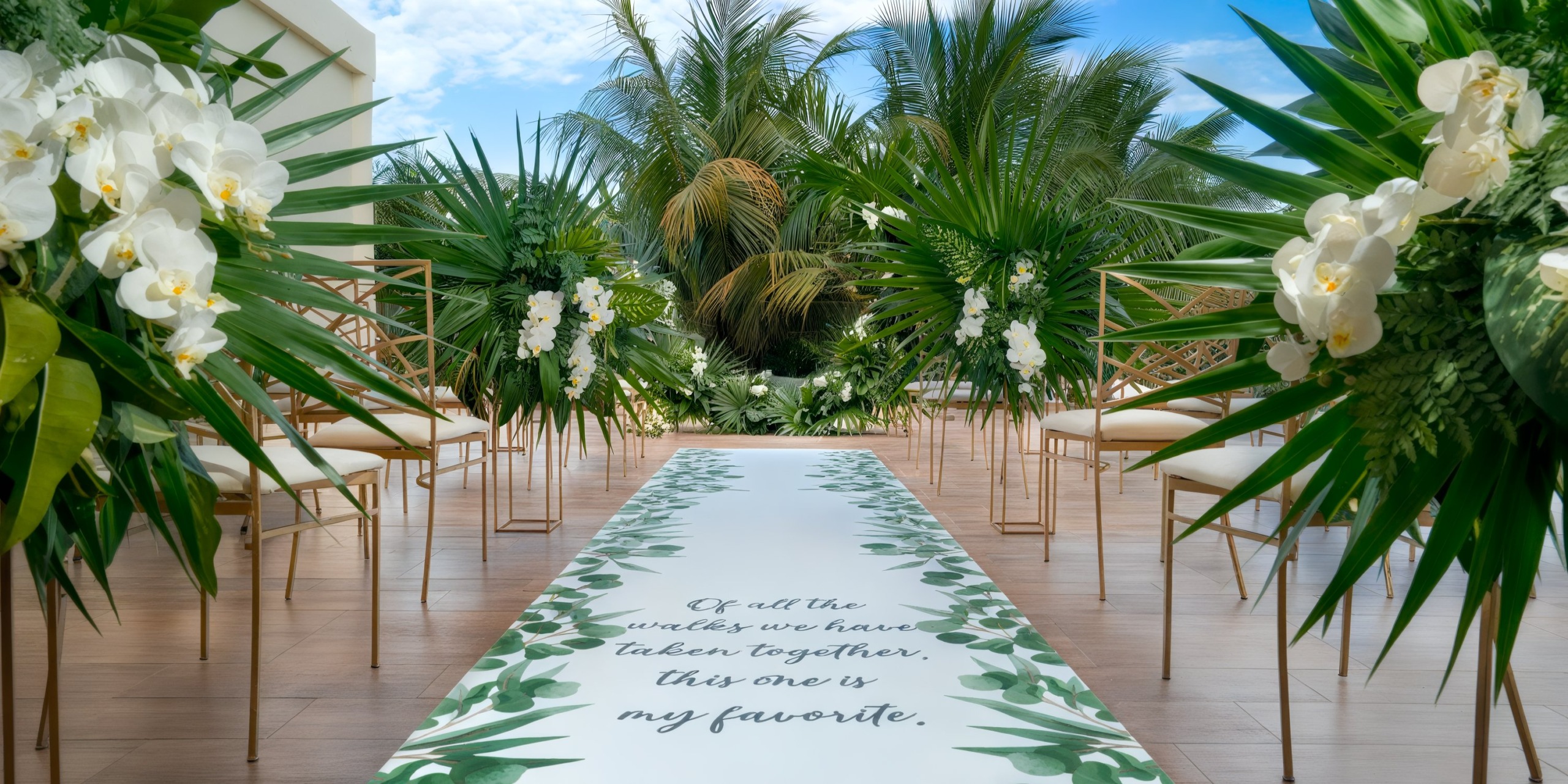 A tropical outdoor wedding at Azul Beach Resort Riviera Cancun features an aisle adorned with white flowers and large green leaves, with a printed romantic message on the runner. Rows of chairs with elegant gold frames flank the aisle.