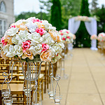 Wedding aisle with pink and white floral arrangements on stands and hanging candles, leading to an arch adorned with similar flowers.