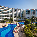 Aerial view of a modern hotel with curved swimming pools, palm trees, and sun loungers, set against a clear blue sky.