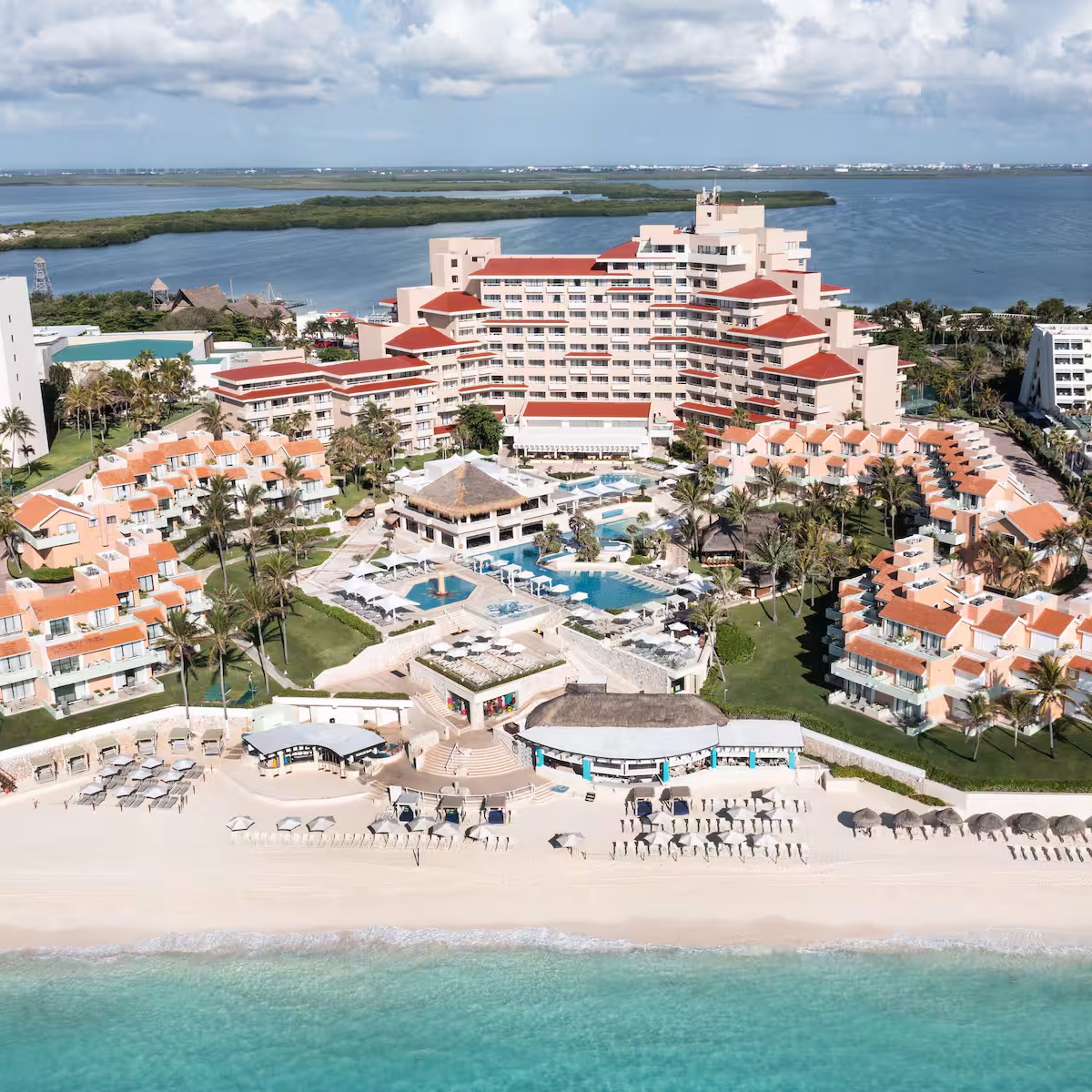 Aerial view of Wyndham Grand Cancun, a beachfront resort featuring multiple pools, red-roofed buildings, and a sandy beach equipped with lounge chairs. The scene is set against the backdrop of the ocean and lush green landscape.