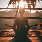 A person sits cross-legged in a meditative pose on a rug outdoors with the sun setting behind palm trees.