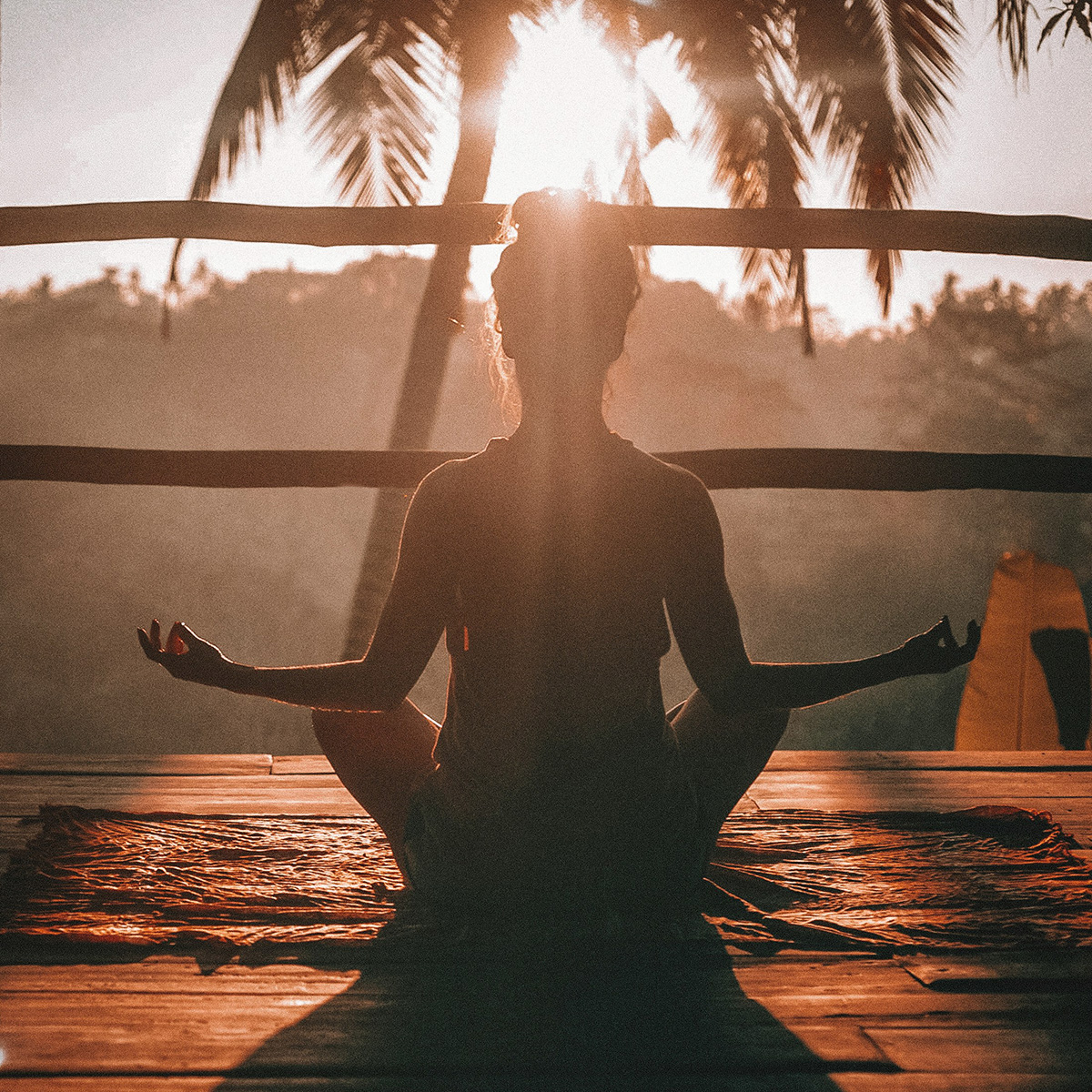 A person sits cross-legged in a meditative pose on a rug outdoors with the sun setting behind palm trees.