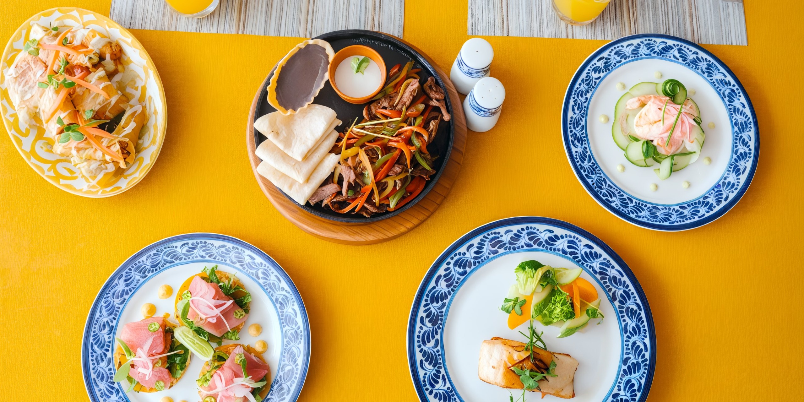 Aerial view of five plates featuring an array of gourmet dishes, offering a diverse selection of meats, vegetables, and sauces, all elegantly arranged on a bright yellow table at Azul Beach Resort Riviera Cancun.