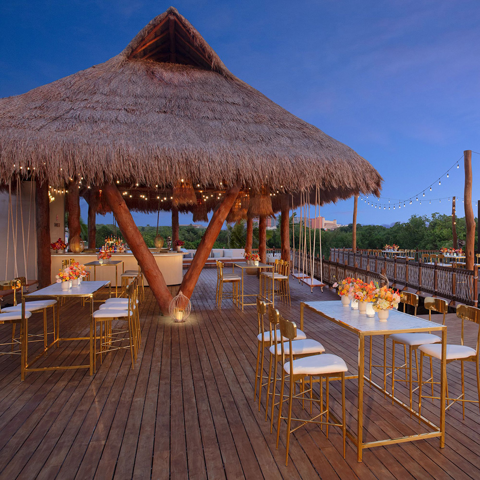 Outdoor venue with a thatched-roof gazebo, decorated with string lights and floral centerpieces. White tables and gold chairs are arranged on a wooden deck.