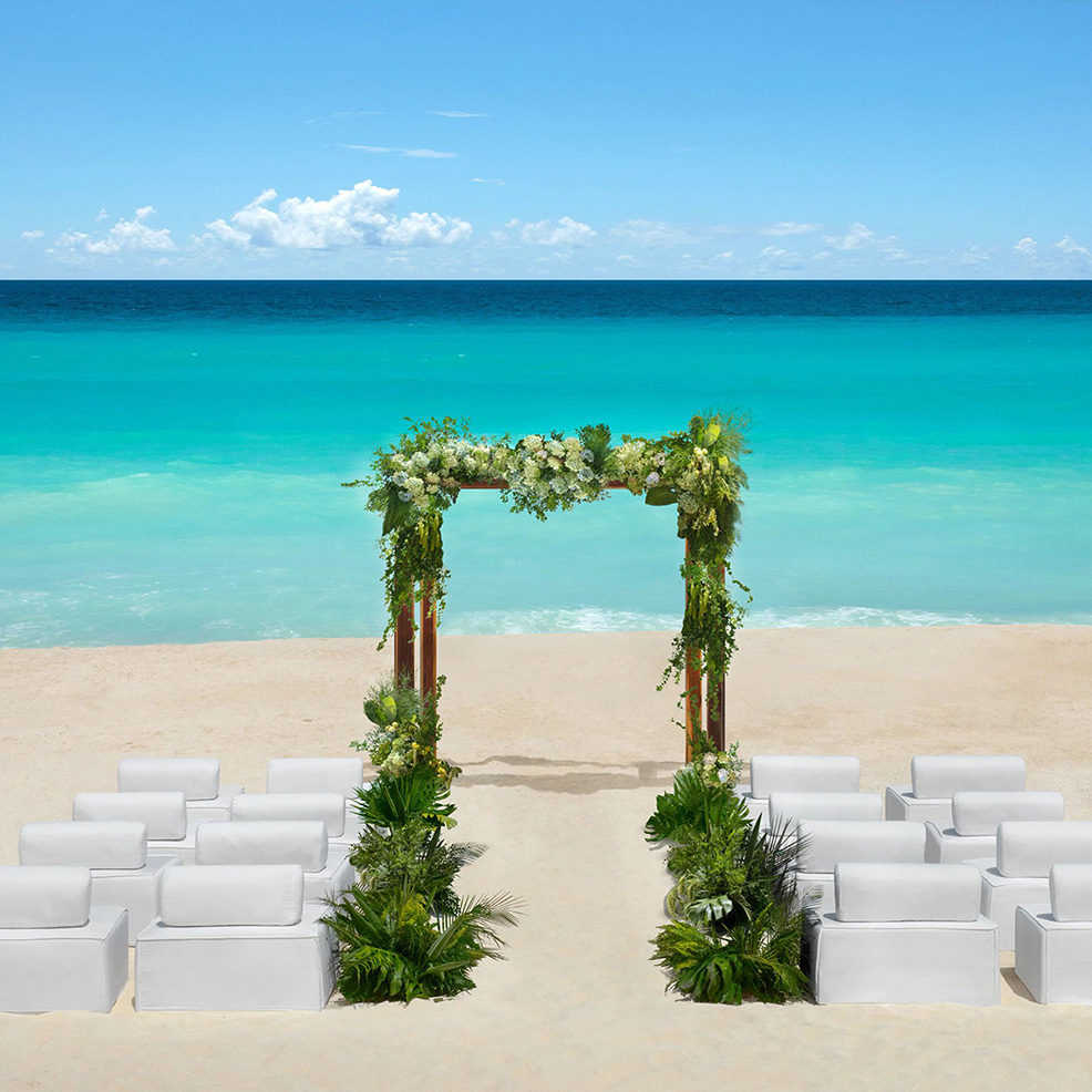 Beach wedding setup with a wooden arch decorated with greenery and white flowers. Rows of white chairs are arranged on the sand, facing the turquoise ocean under a clear sky.