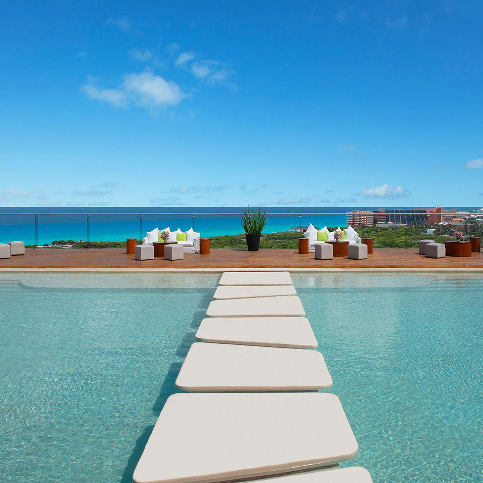 A swimming pool with a floating stepping stone path overlooks a clear blue ocean, amid modern outdoor seating and a distant resort view.
