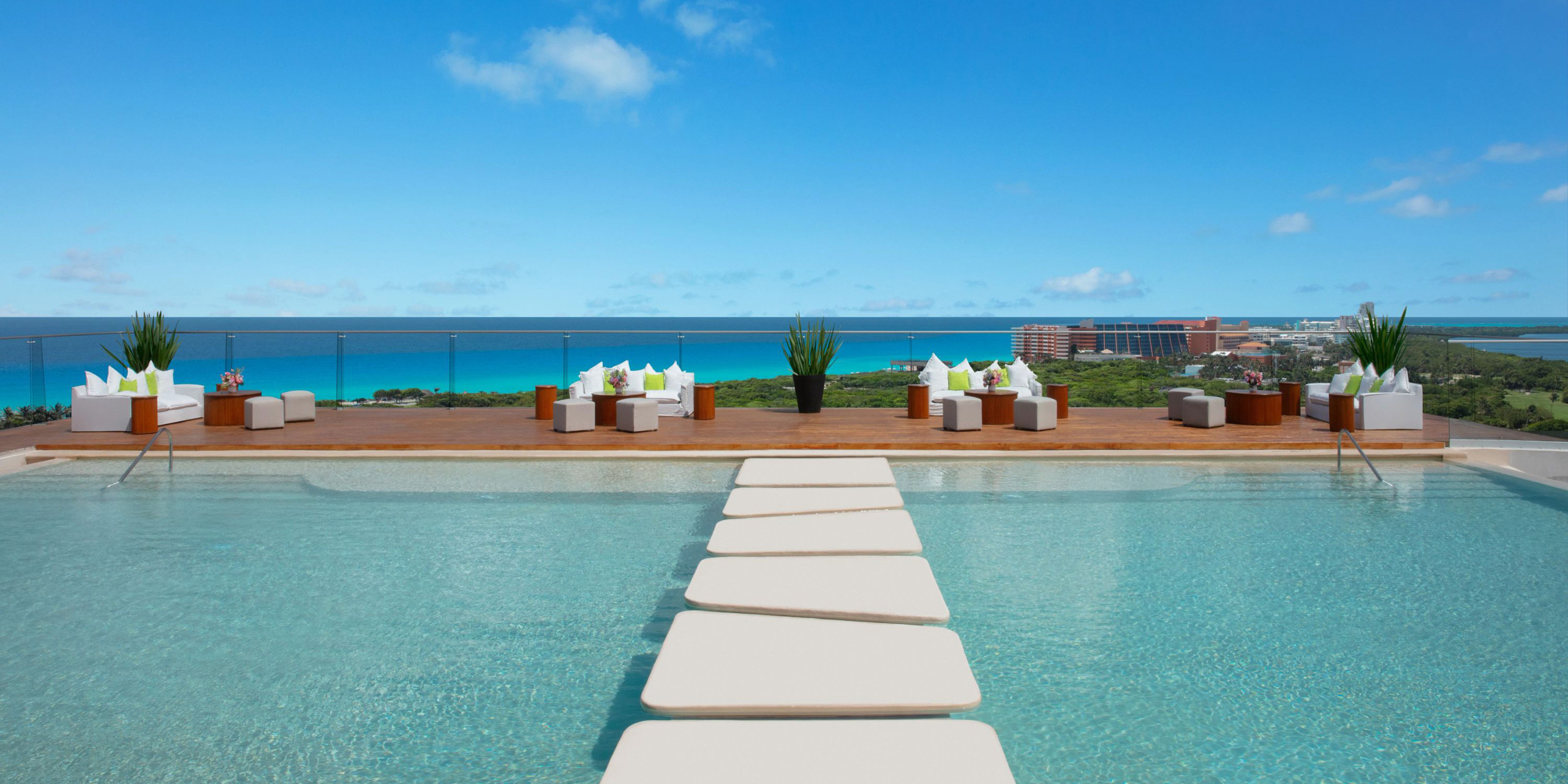 The Hyatt Vivid Grand Island features a rooftop pool with stepping stones that lead to a cozy seating area, with breathtaking views of the ocean and cityscape against a clear blue sky.