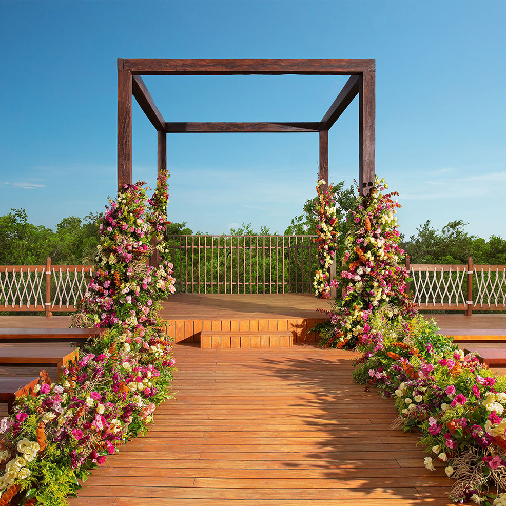 A wooden outdoor aisle leads to a square arch adorned with colorful flowers, set against a backdrop of greenery and a clear blue sky.