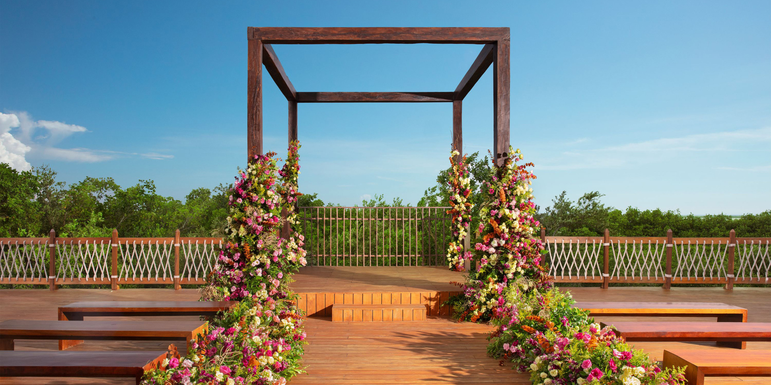 Set against a backdrop of greenery and a clear blue sky, the Hyatt Vivid Grand Island features an outdoor wedding altar with a wooden frame adorned with floral decorations, surrounded by benches on a wooden deck.