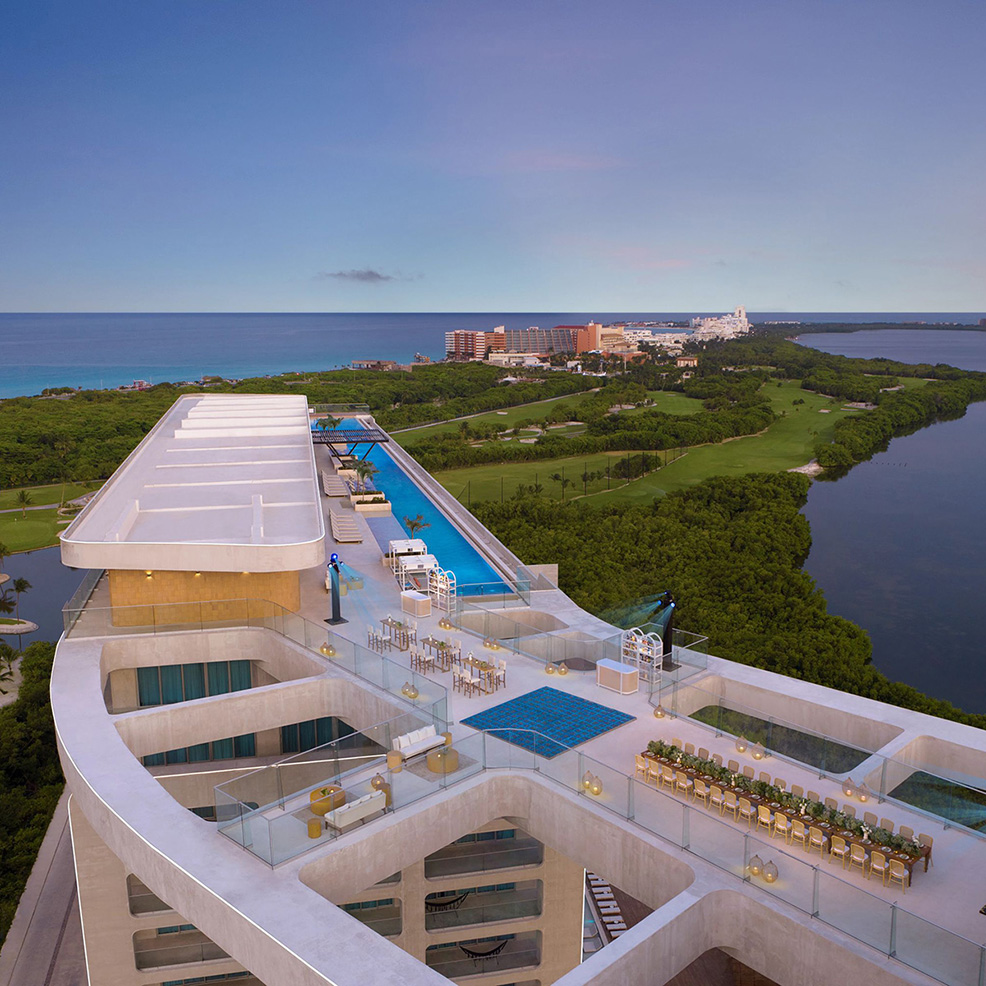 Rooftop infinity pool and terrace overlooking a coastal cityscape, with ocean and greenery in the background.