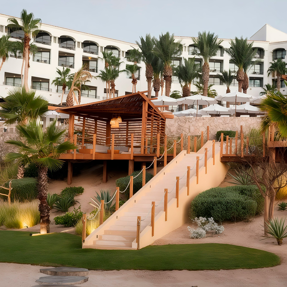 The Hilton Los Cabos features an outdoor wooden gazebo and staircase surrounded by palm trees and lush greenery, with a white building in the background.