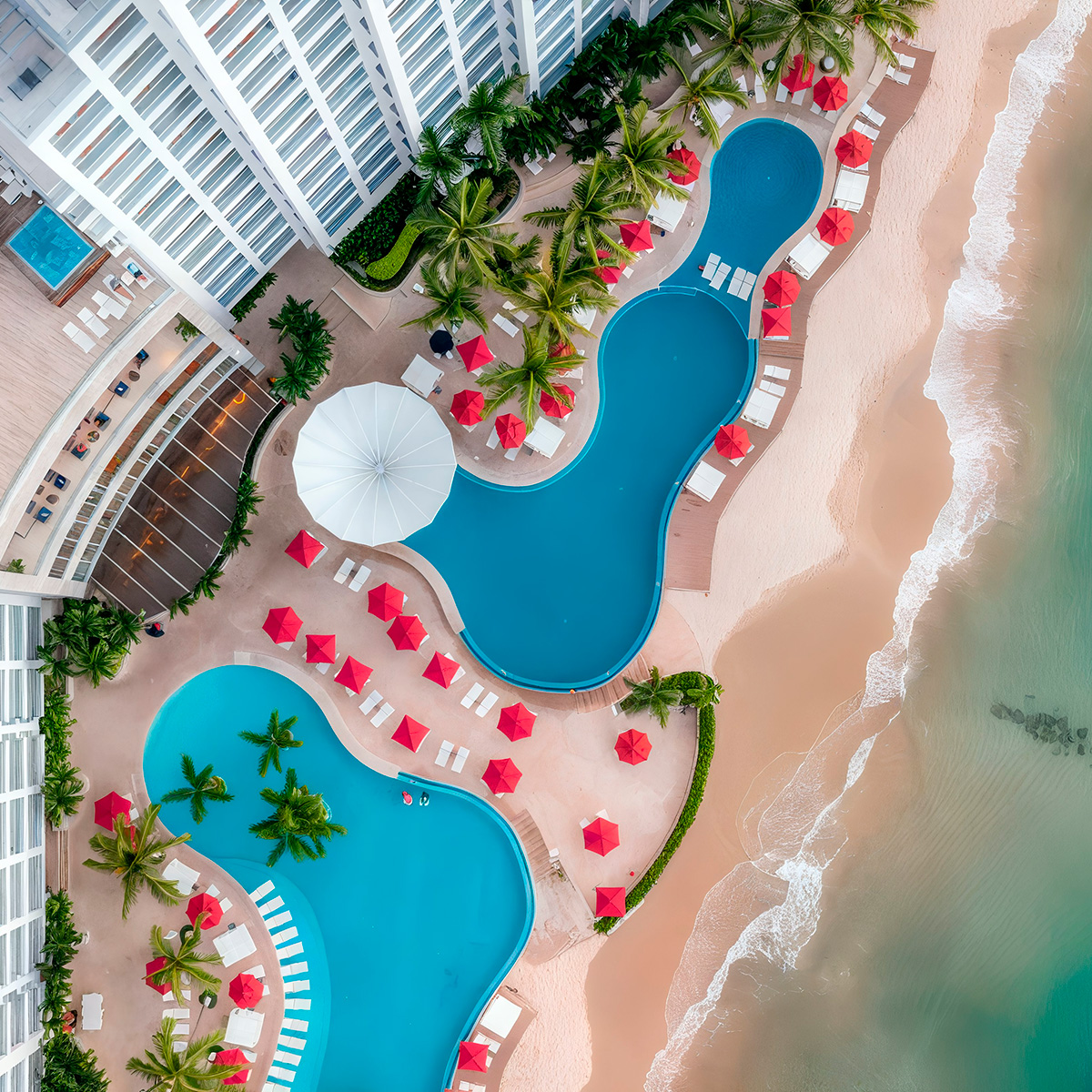 Aerial view of the Hilton Vallarta Riviera All-Inclusive Resort with two irregularly shaped pools bordered by red umbrellas and palm trees, next to a sandy beach and ocean.