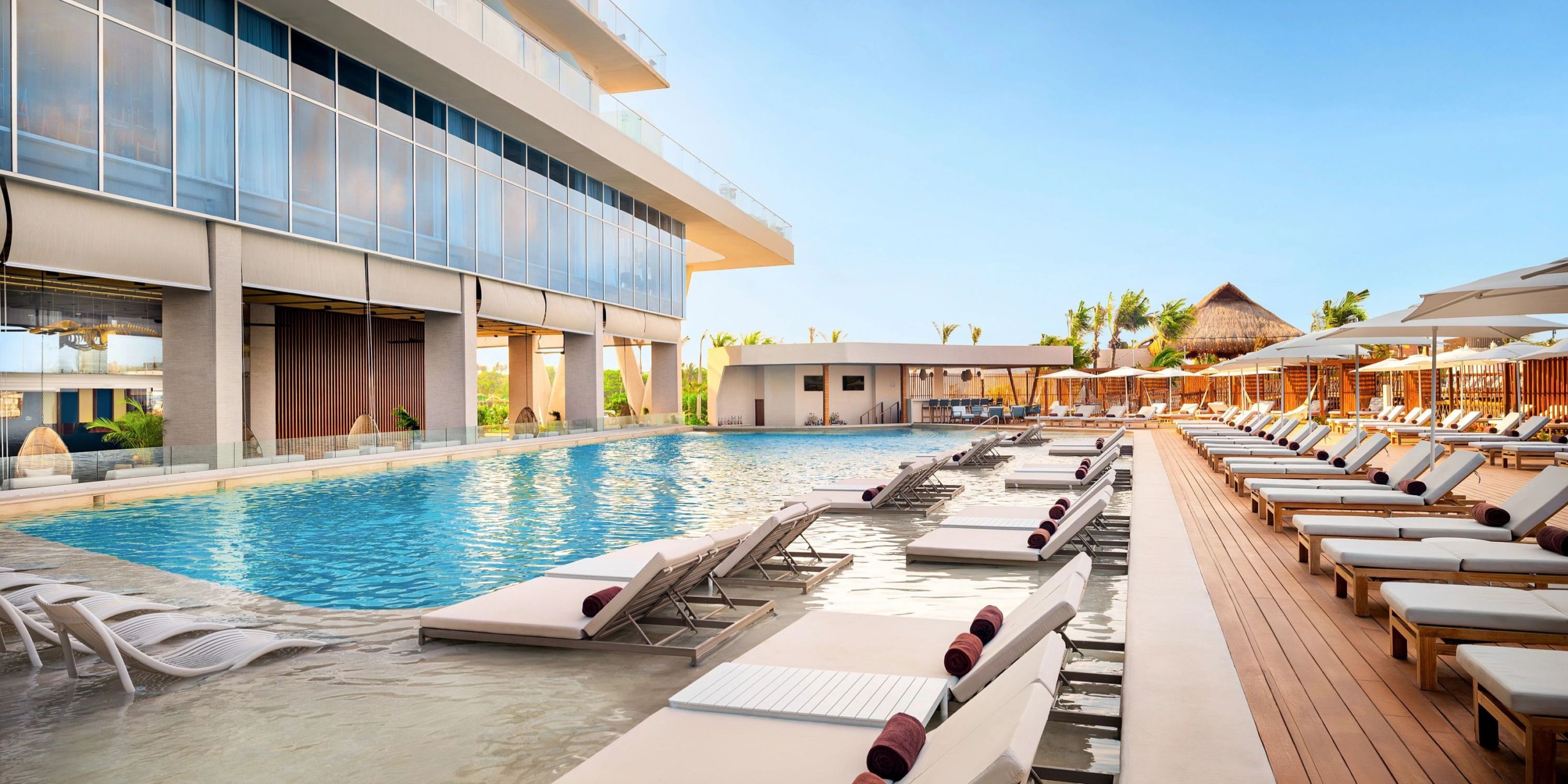 The Hyatt Vivid Grand Island features an outdoor pool area complete with lounge chairs and sun umbrellas lining the deck. In the background, a modern building stands tall alongside palm trees, all set under a clear blue sky.