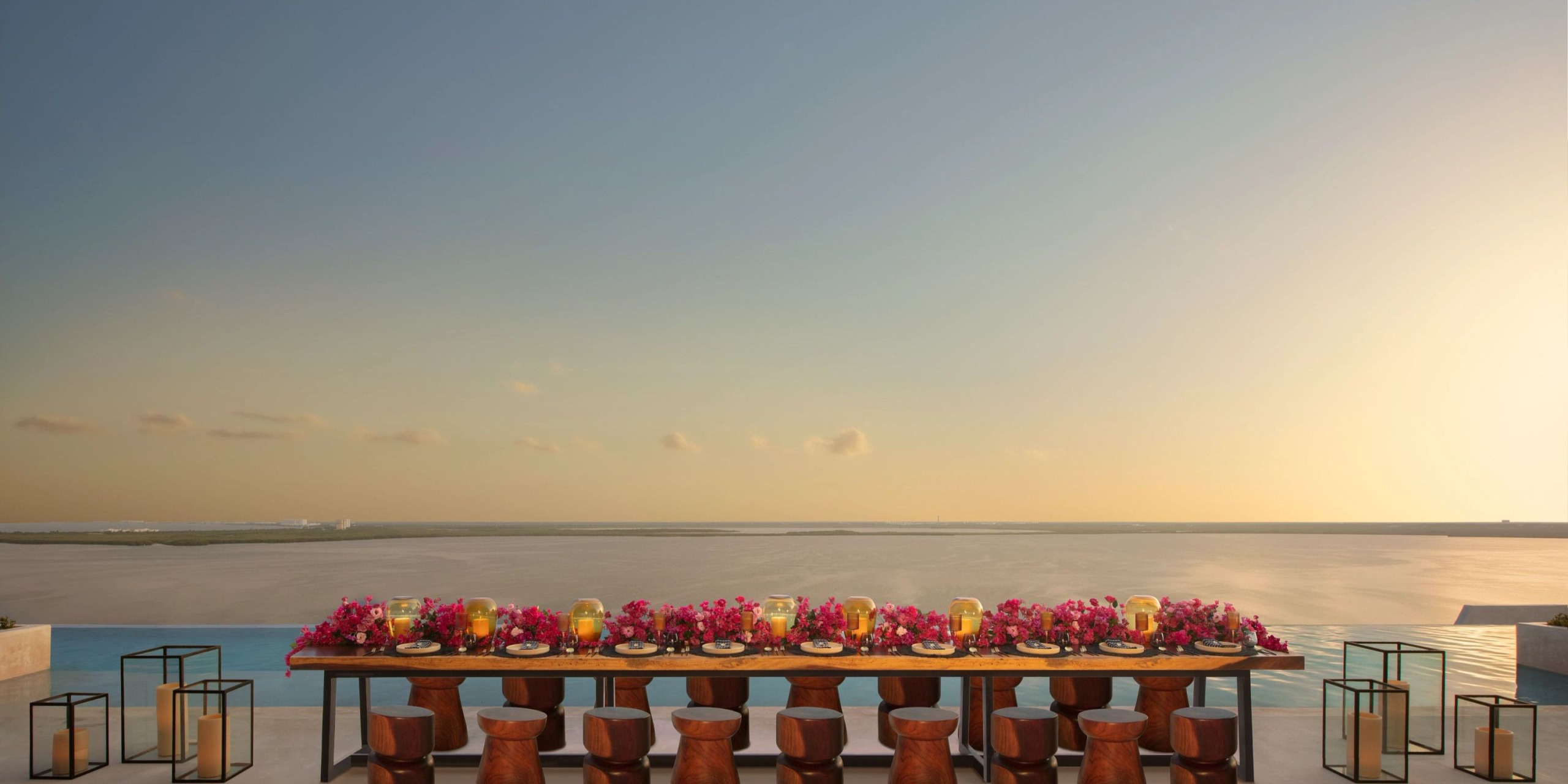 A dining setup at the Hyatt Vivid Grand Island, adorned with pink flowers and candles, overlooks a tranquil body of water beneath a clear sunset sky.