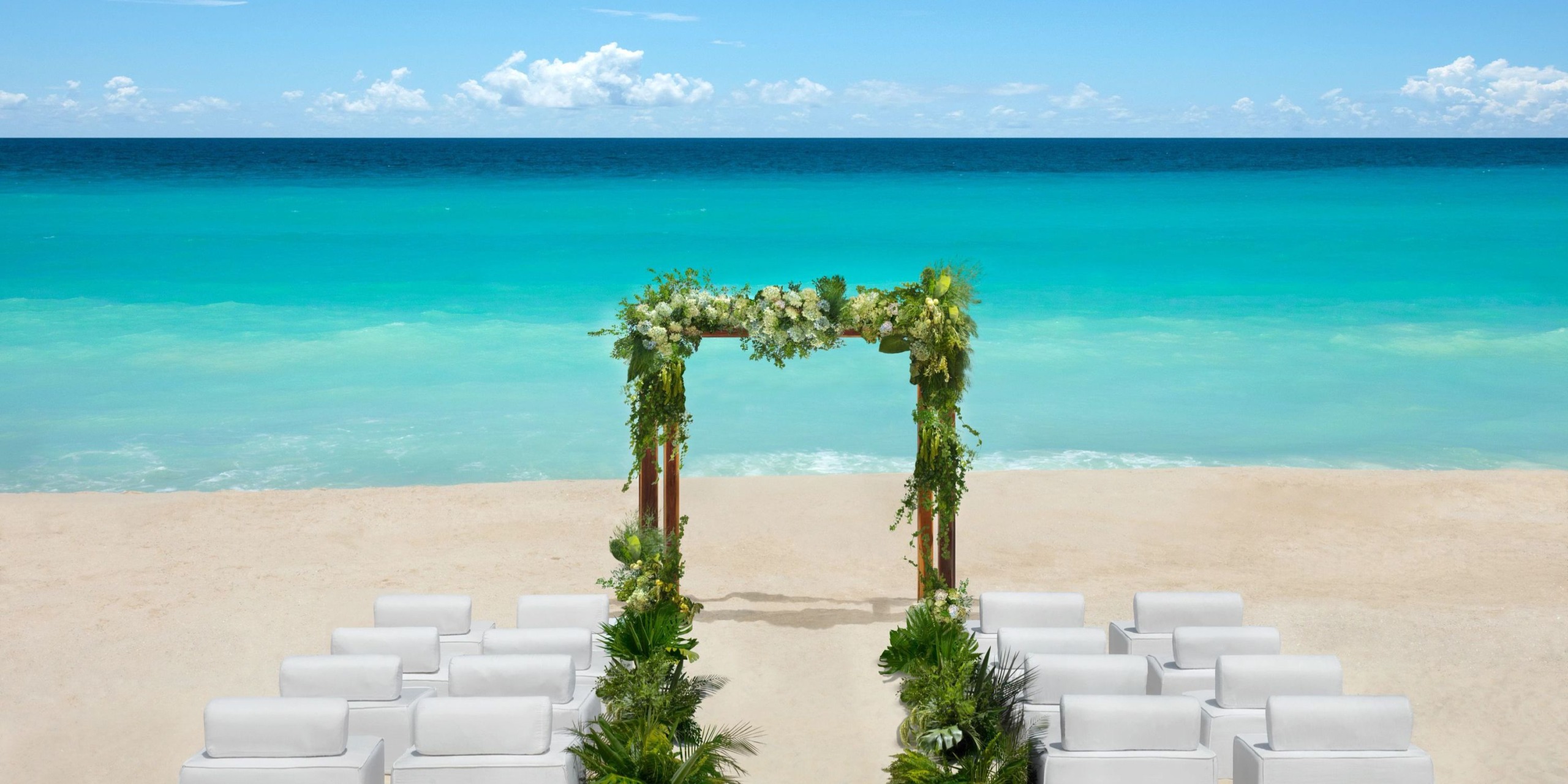 Experience a picturesque beach wedding at Hyatt Vivid Grand Island, featuring a stunning floral arch and elegant white chairs, all set against the backdrop of the ocean under a clear blue sky.